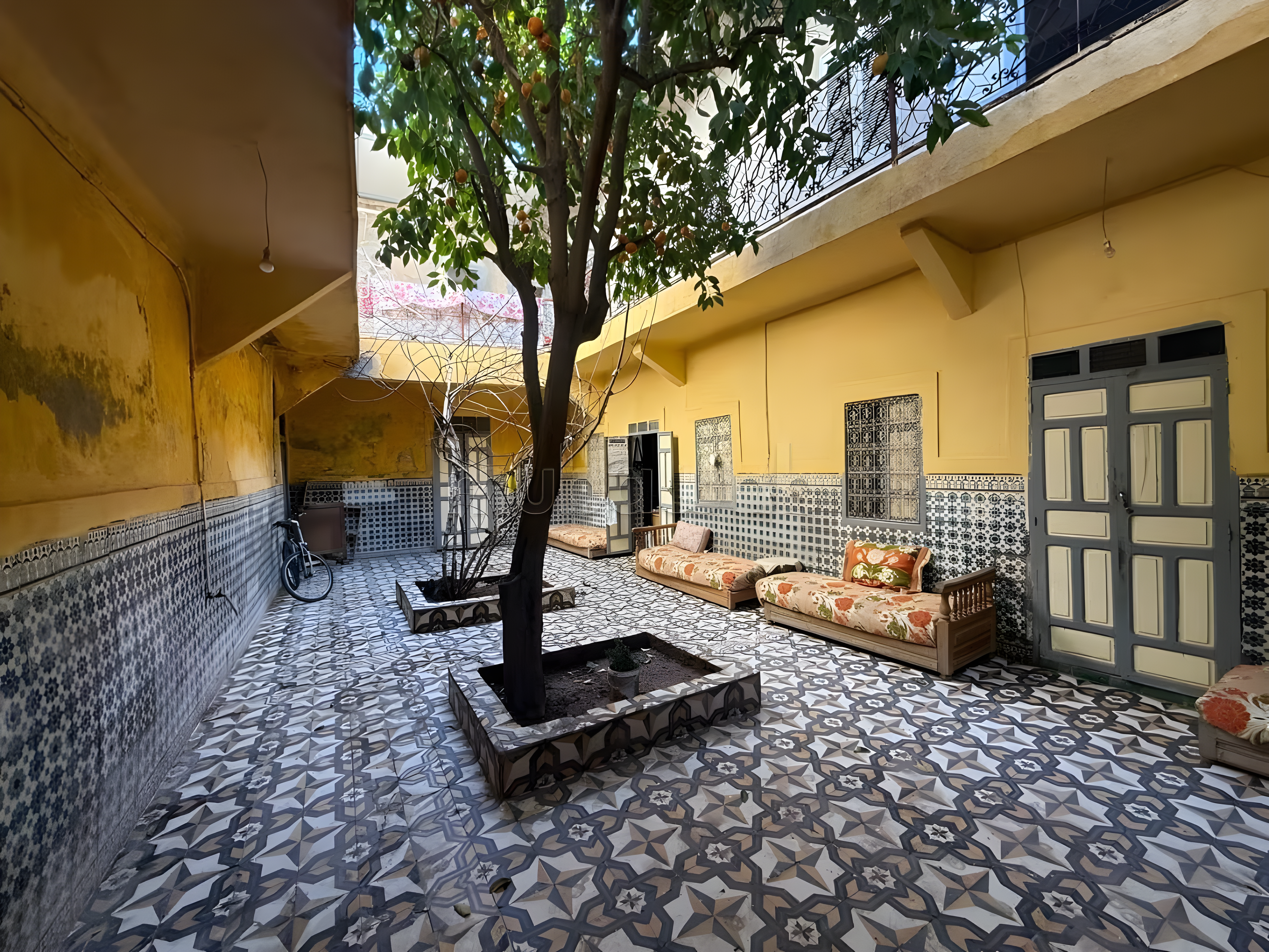 Central courtyard with mosaic tiles, yellow walls, tree, and seating in Moulay Lyazid guesthouse.