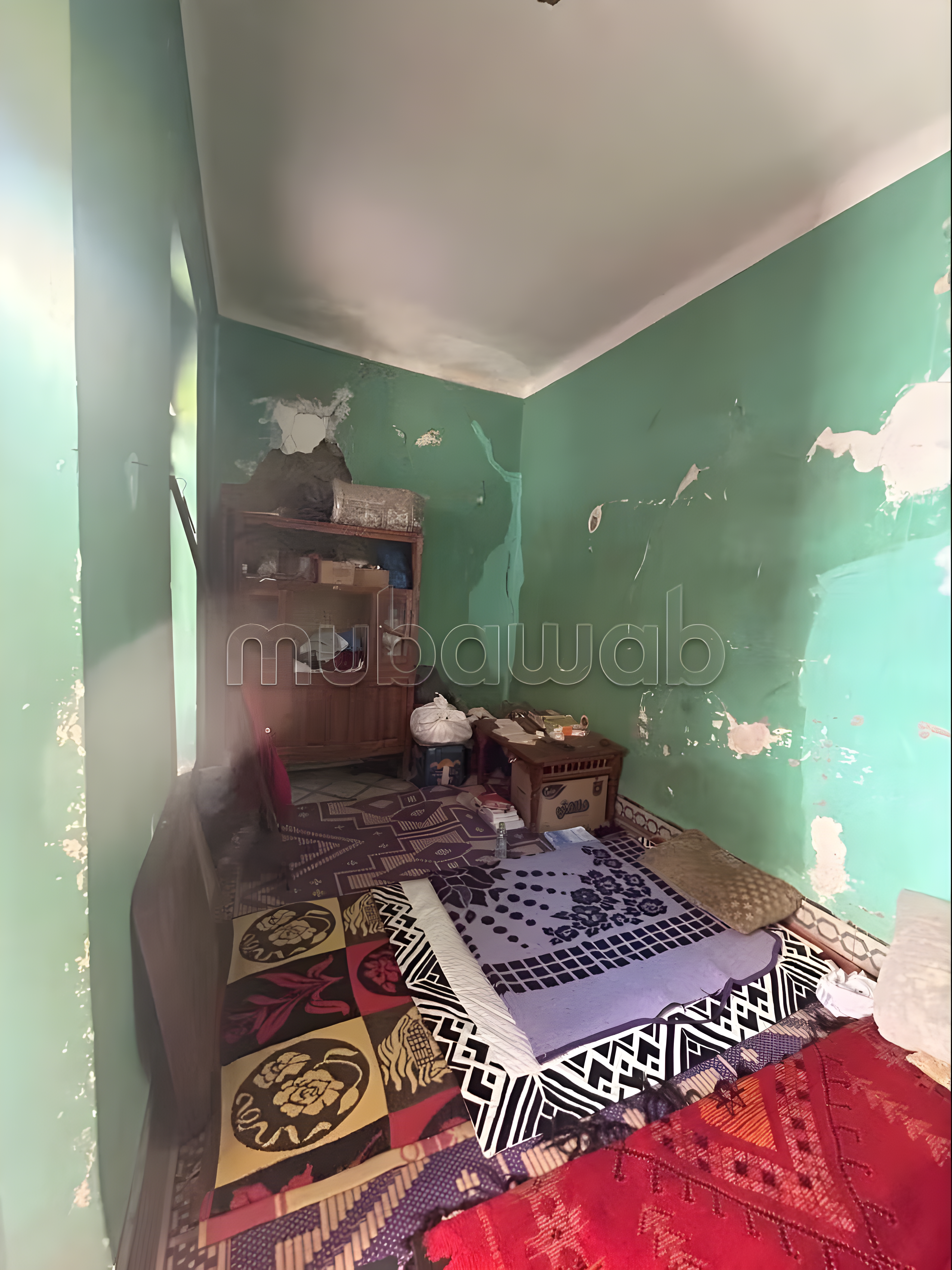Small Kennaria riad bedroom with green walls, peeling paint, wooden shelf, and layered rugs.